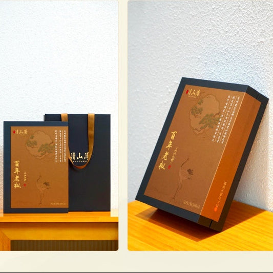 A boxed product set shown in two clean studio-style arrangements: a smaller matching box and dark tote bag on a wooden surface against a light wall, and a single tall rectangular box standing upright on the right. The packaging is predominantly warm brown/tan with black side panels and a small gold handle on the bag.